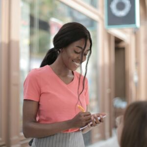 Smiling waitress in a pink shirt taking an order at an outdoor cafe.